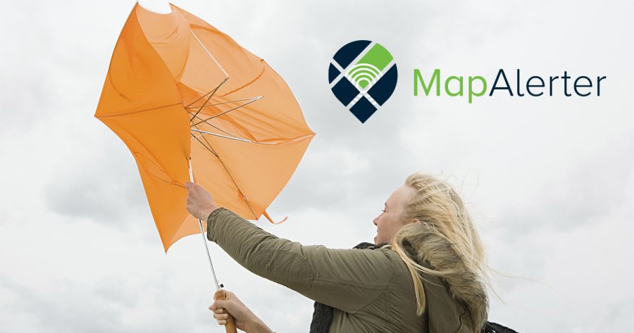 A woman struggles against strong wind holding an orange umbrella that has flipped inside out. She is wearing a green parka with a fur-lined hood and a black scarf, with her blonde hair blowing in the wind against a cloudy sky.